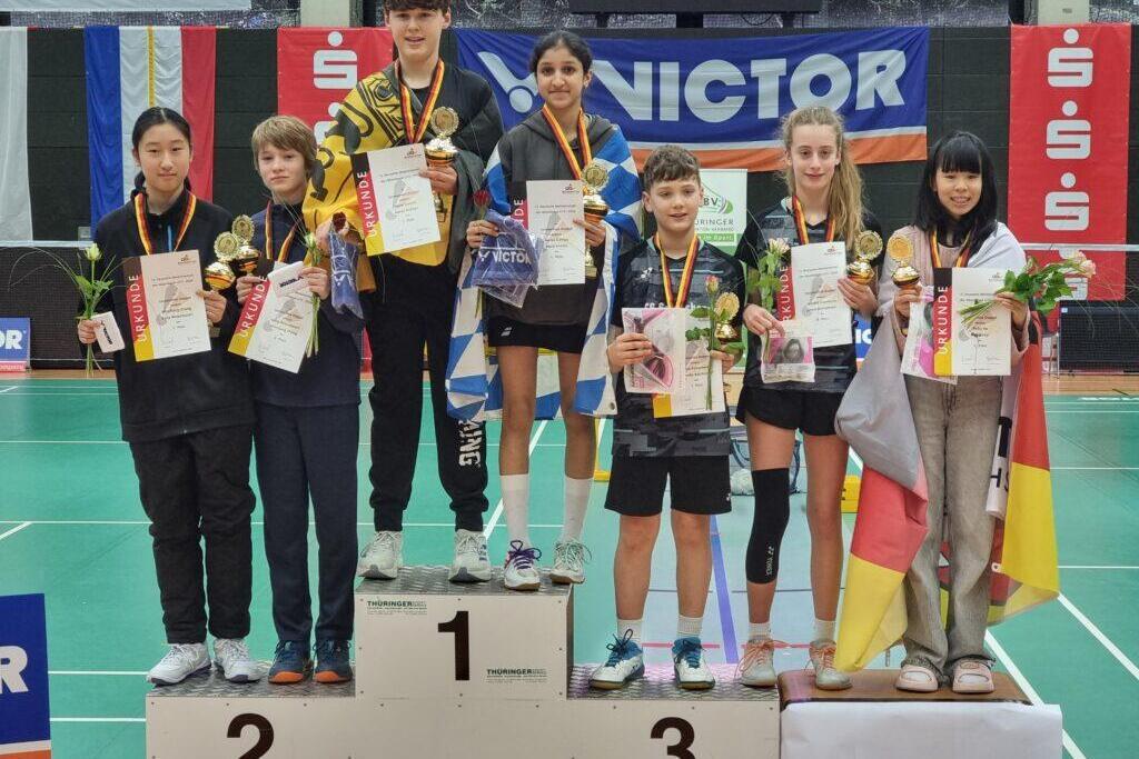 Drei Jungen und vier Mädchen stehen auf einem Siegertreppchen, halten Urkunden und Medaillen in einer Sporthalle.