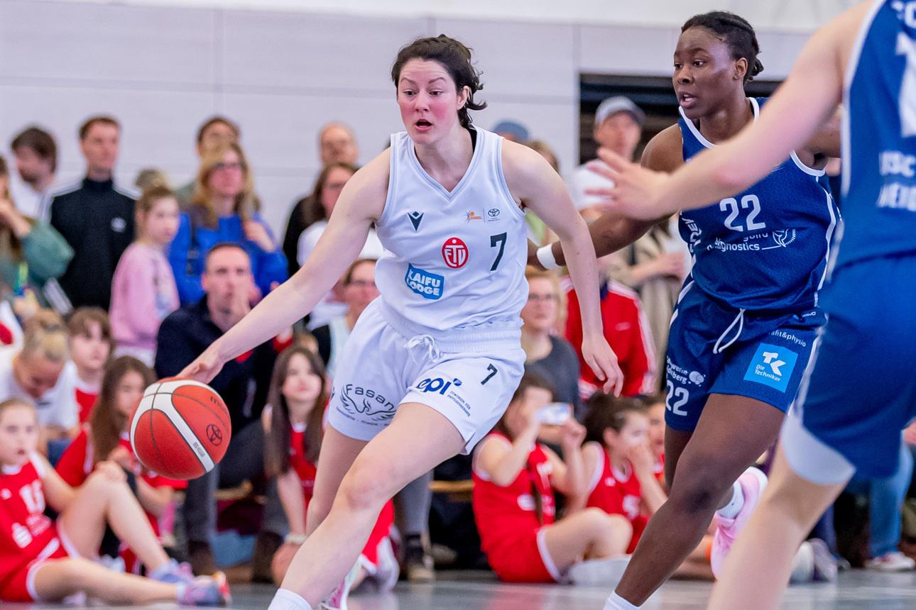 Basketballspiel mit zwei Spielerinnen im Wettkampf; eine dribbelt den Ball, während die andere verteidigt. Zuschauer im Hintergrund.