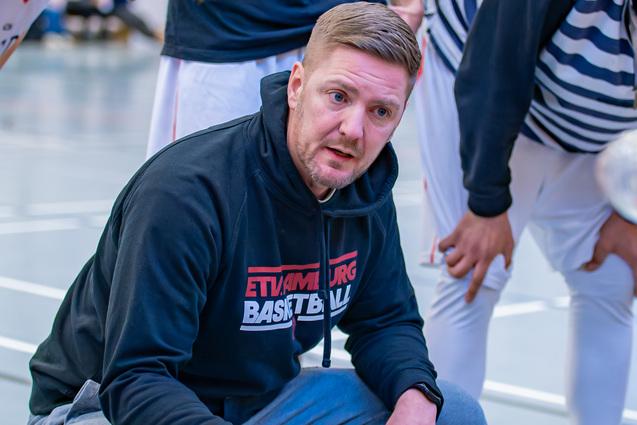 Basketballtrainer kniet im Gespräch mit Spielern, während sie sich um ihn versammeln und Waterflaschen halten.