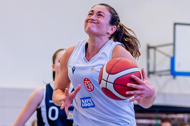 Basketballspielerin mit Ball in der Hand springt zum Korb, während eine Gegnerin sie verfolgt. Zuschauer im Hintergrund.