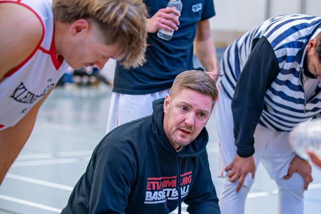Basketballtrainer kniet im Gespräch mit Spielern, während sie sich um ihn versammeln und Waterflaschen halten.