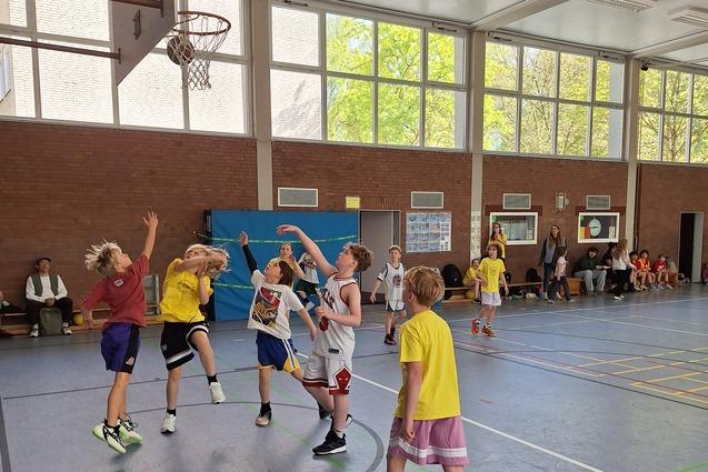 Mehrere Kinder spielen Basketball in einer Halle, während Zuschauer auf Bänken am Rand sitzen.