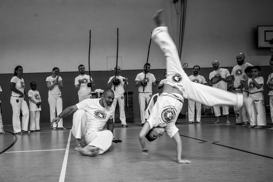 Zwei Personen in weißen Trainingsanzügen führen eine Bewegung im Capoeira vor, umgeben von Zuschauern in einem Sportraum.