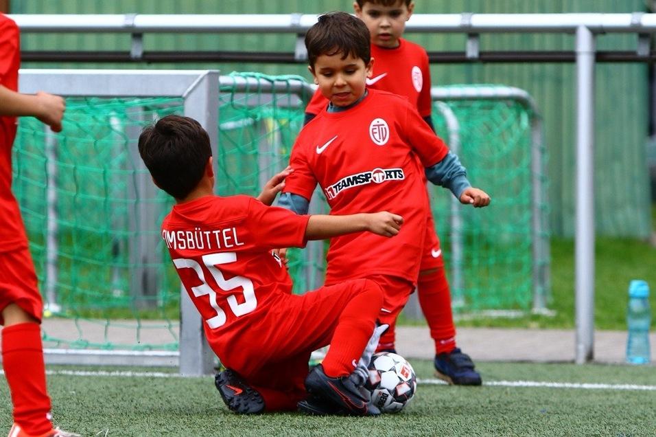 Zwei Kinder in roten Fußballtrikots spielen auf einem Rasenplatz; einer sitzt und greift den anderen an.