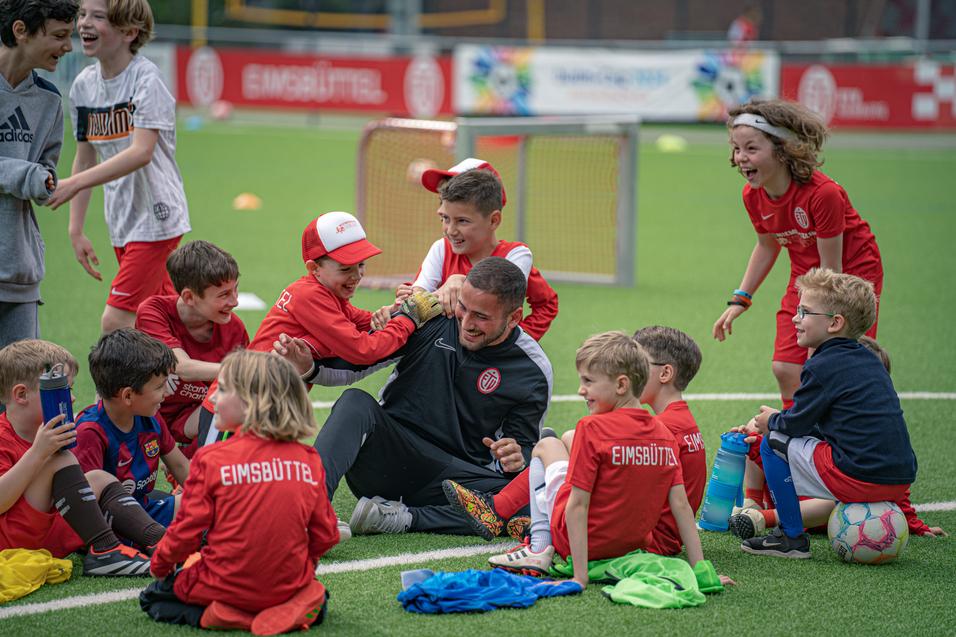 Gruppe von Kindern in Fußballtrikots, die um einen Trainer auf dem Kunstrasen lachen und spielen.
