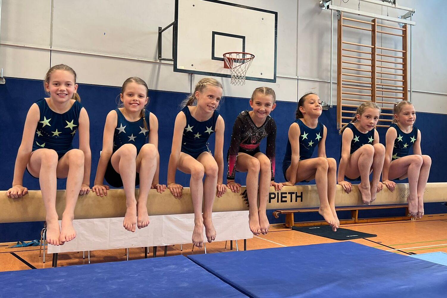 Gruppe von sieben Mädchen in Sportanzügen sitzt auf einem Schwebebalken in einer Turnhalle.