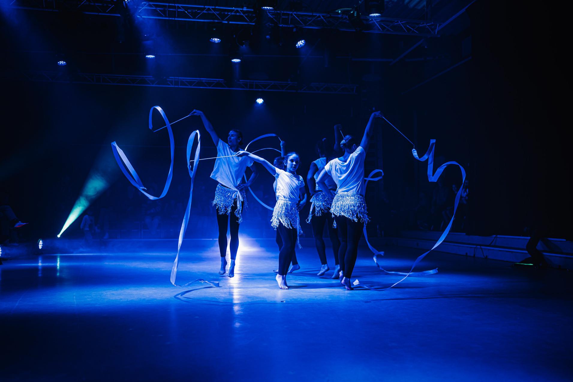Tänzerinnen in weißen Oberteilen und schwarzen Leggings führen eine Choreografie mit farbigen Bändern auf einer Bühne auf.