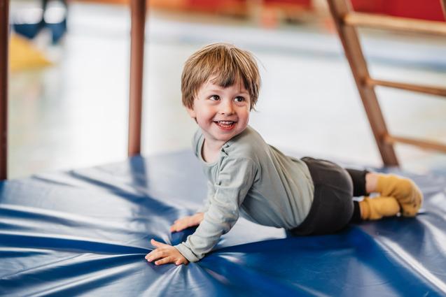 Lächelnder Junge liegt auf einer blauen Gymnastikmatte und spielt in einer Turnhalle.