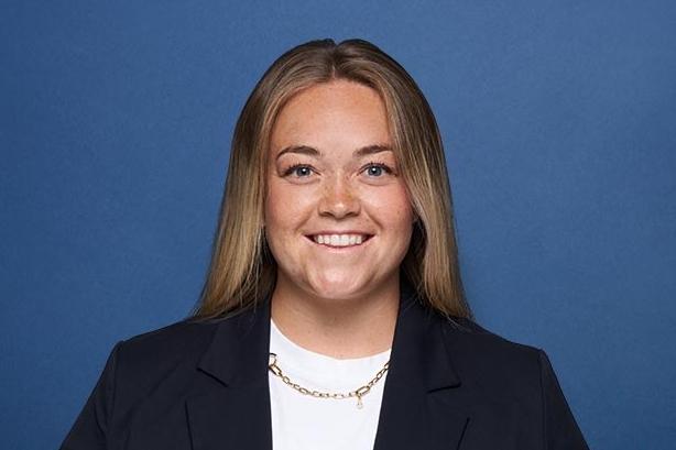 Portrait einer lächelnden Frau mit langen Haaren, in einem dunklen Blazer und weißem Oberteil, vor blauem Hintergrund.
