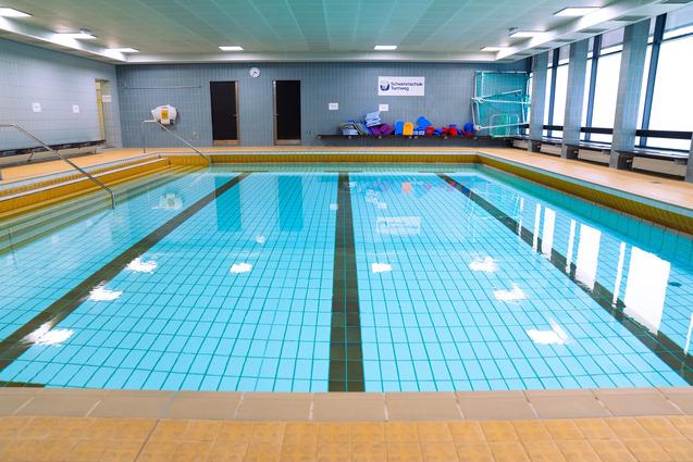 Innenansicht eines leeren Schwimmbeckens mit Fliesen und Treppen, umgeben von Fenstern und Bänken.