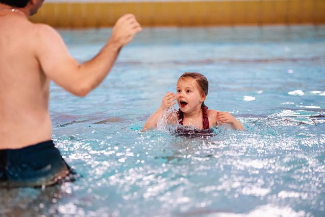 Ein Erwachsenen und ein aufgeregtes Kind spielen im Wasser eines Schwimmbeckens. Spritzer sind sichtbar.