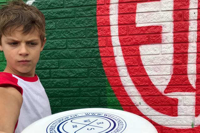 Ein Junge hält eine Frisbee in Richtung Kamera, vor einer bunten Wand mit grünen und roten Mustern.