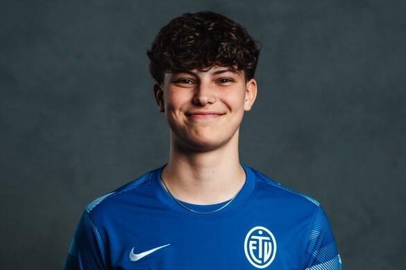 Porträt eines lächelnden jungen Sportlers in blauer Teamkleidung mit Logo, vor einem neutralen Hintergrund.